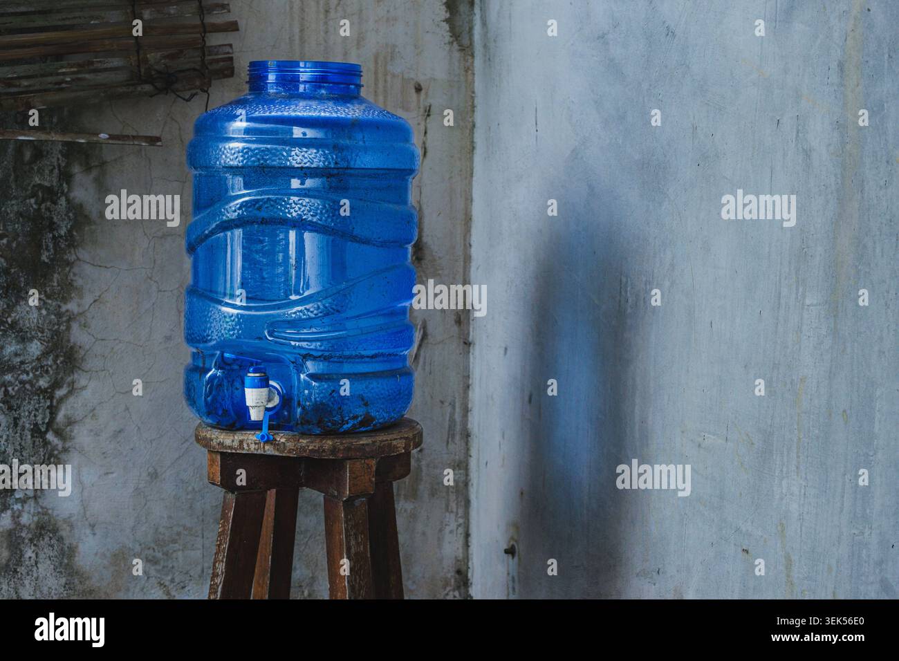 Eine blaue, durchscheinende Wasserspenderflasche aus Kunststoff mit Wasserhahn, die auf einem verwitterten Holzhocker gegen eine rustikale Betonwand gestellt wird. Stockfoto