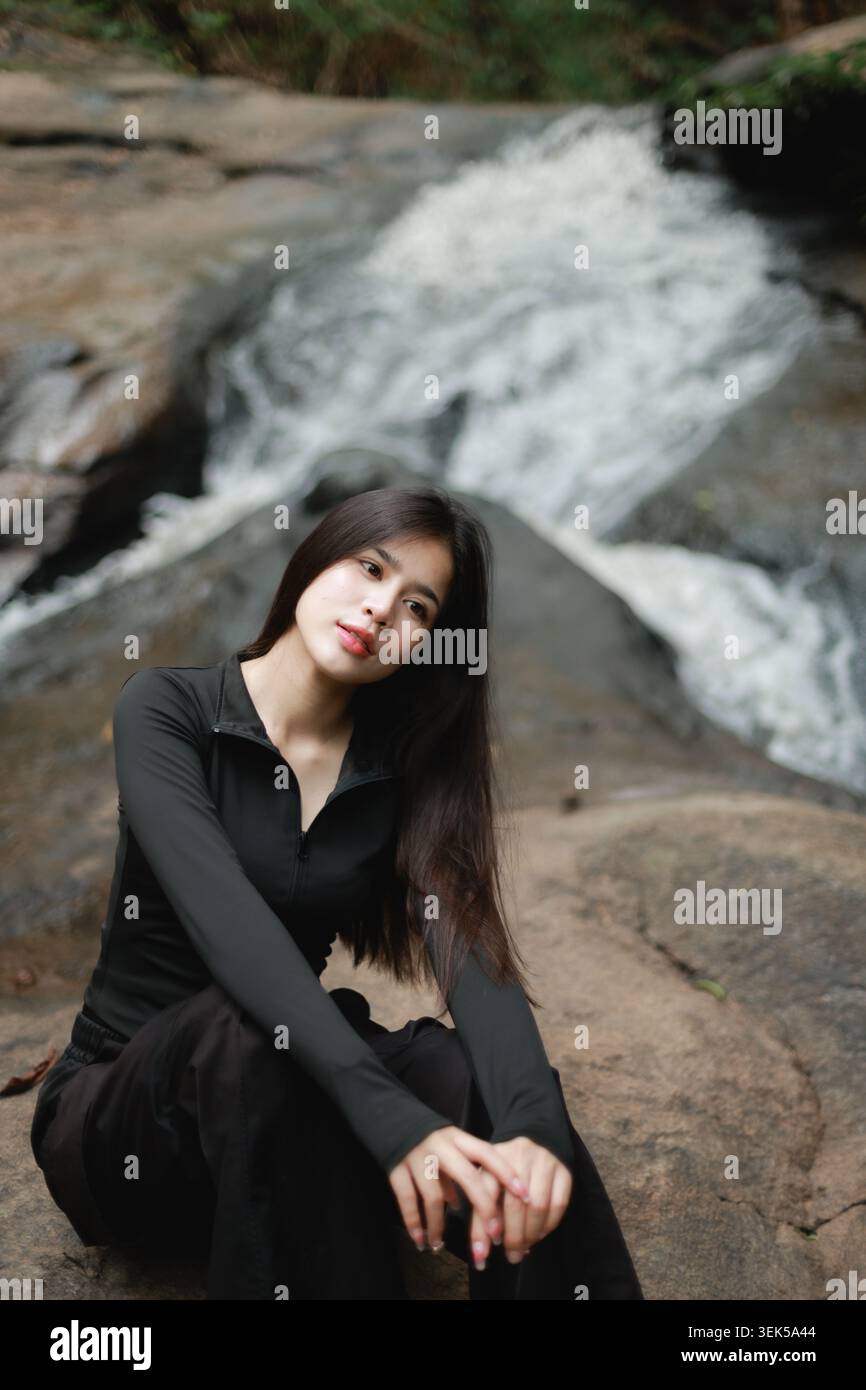 Wunderschöne junge asiatische Frau, die dunkelbrauner Felsen am fließenden Wasserfall sitzt, natürliche Waldumgebung, die ruhig und friedlich mit ruhiger, selbstbewusster Ex aussieht Stockfoto