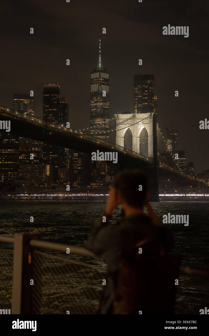 Verschwommene Person, die nachts die brooklyn Bridge und die Skyline von manhattan vom Ufer aus fotografiert. Stockfoto
