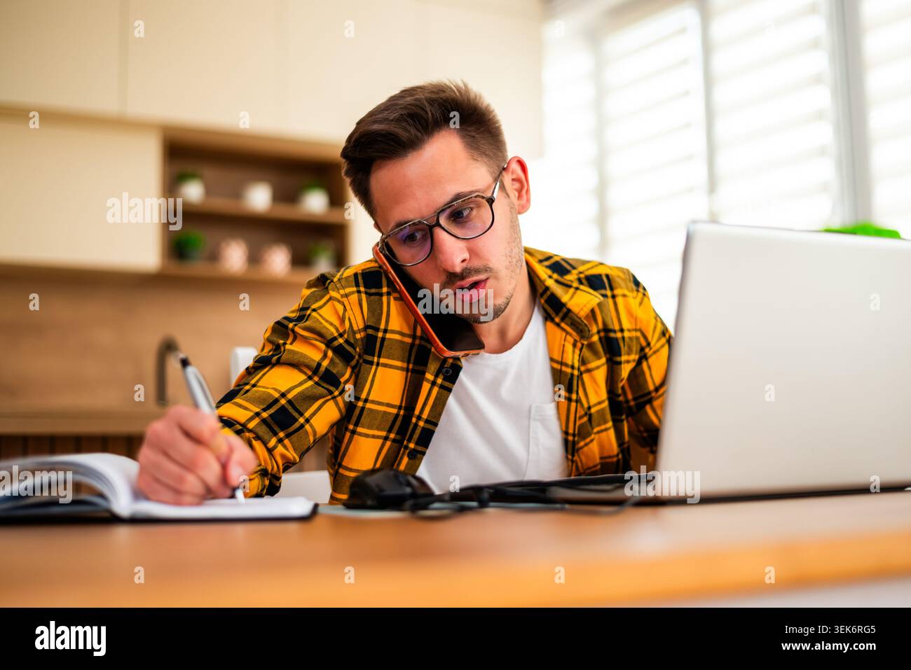 Ein junger Mann arbeitet von zu Hause aus, um Ideen und Erinnerungen aufzuschreiben und dabei Online-Forschung mit traditionellem Notizen zu verbinden. Stockfoto
