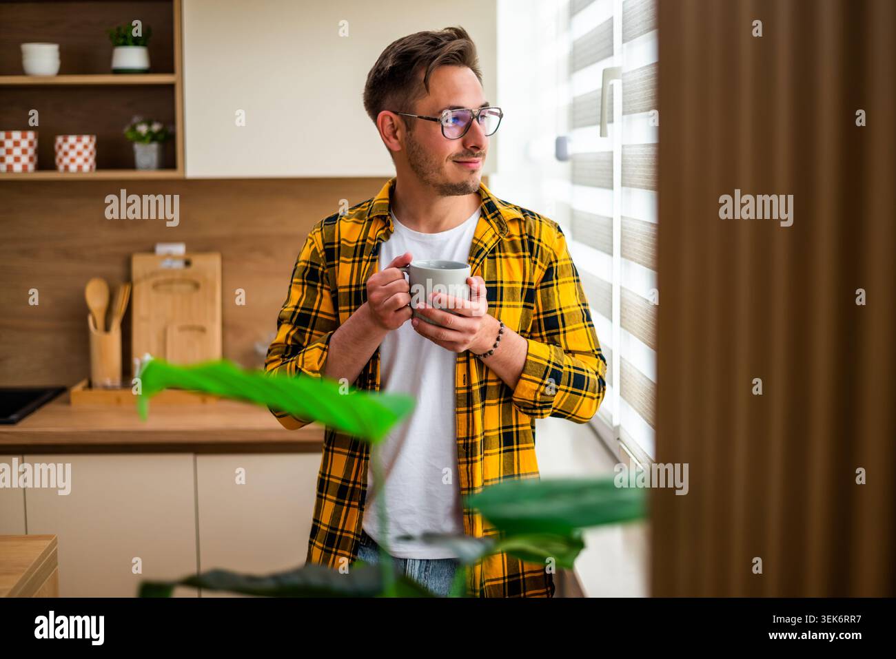 Der junge Mann steht am Fenster in seiner Küche und trinkt Kaffee. Er schaut nach draußen. Stockfoto