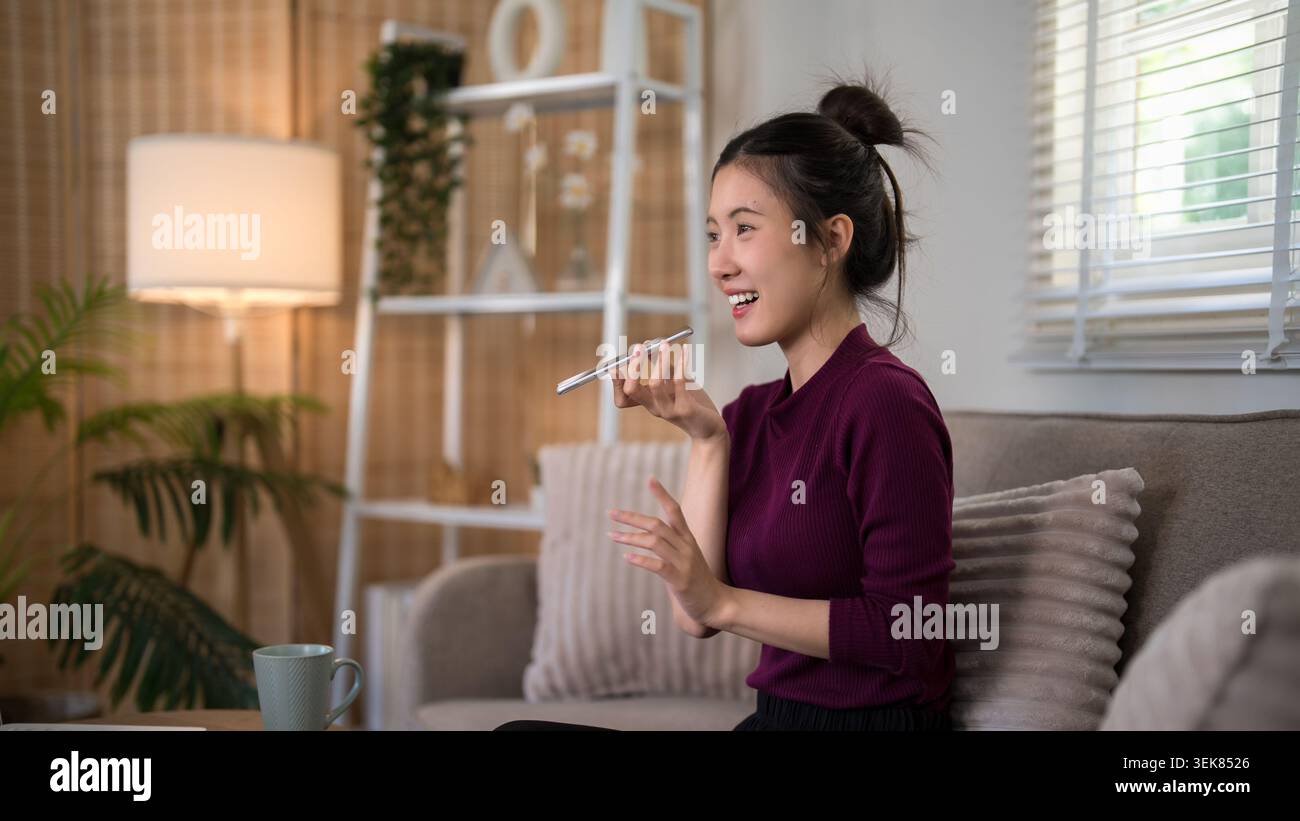 Asiatische Frau professionell sprechendes Smartphone Sprachnachricht Fernarbeit Home Office komfortable moderne Wohnzimmerumgebung Stockfoto