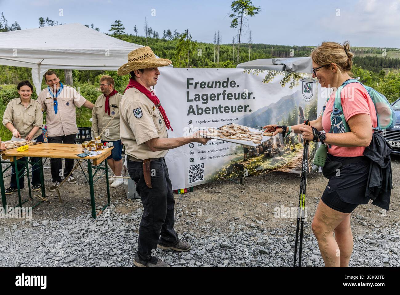 Ein Pfadfinder in Uniformhänden ein Teilnehmer am 14. Frankenwald Wandermarathon ein Stück Honigbrot aus einem Tablett – im Hintergrund steht das Banner der Pfadfindertruppe mit dem Slogan „Freunde“. Lagerfeuer. Abenteuer.“, Ludwigsstadt, Bayern, Deutschland Stockfoto
