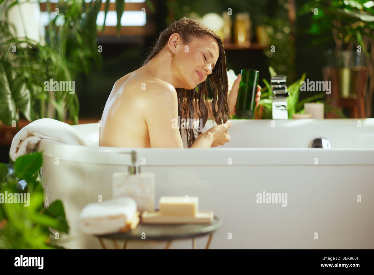 Frau mittleren Alters mit nassbraunem Haar entspannt sich in der weißen Badewanne, Augen geschlossen, Produkt aus dunkelgrüner Flasche auftragen. Grüne Pflanzen und weiche Beleuchtung cre Stockfoto