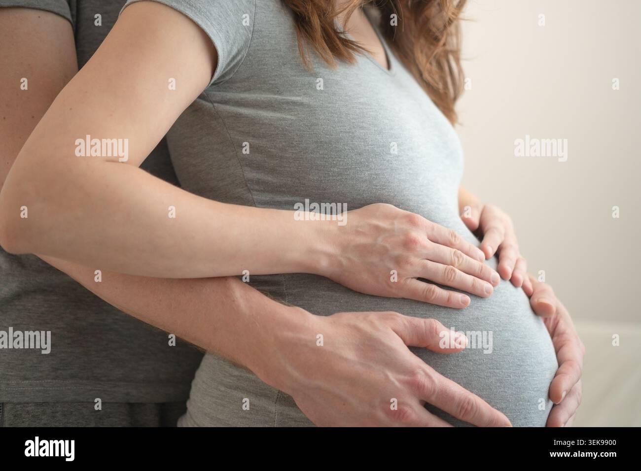 Schwangere Frau mit Partner, der im gemeinsamen intimen Moment die Hände über den Bauch legt. Erwartende Mutter und Ehemann, die sich von Side im Studio mit neutralem Hintergrund umarmen. Werdende Mutter feiert gemeinsam Stockfoto