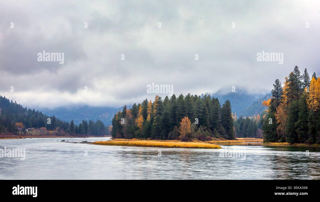 Der Fluss schlängelt sich durch eine ruhige Wildnis mit Herbstbäumen und bewölkten Bergen Stockfoto