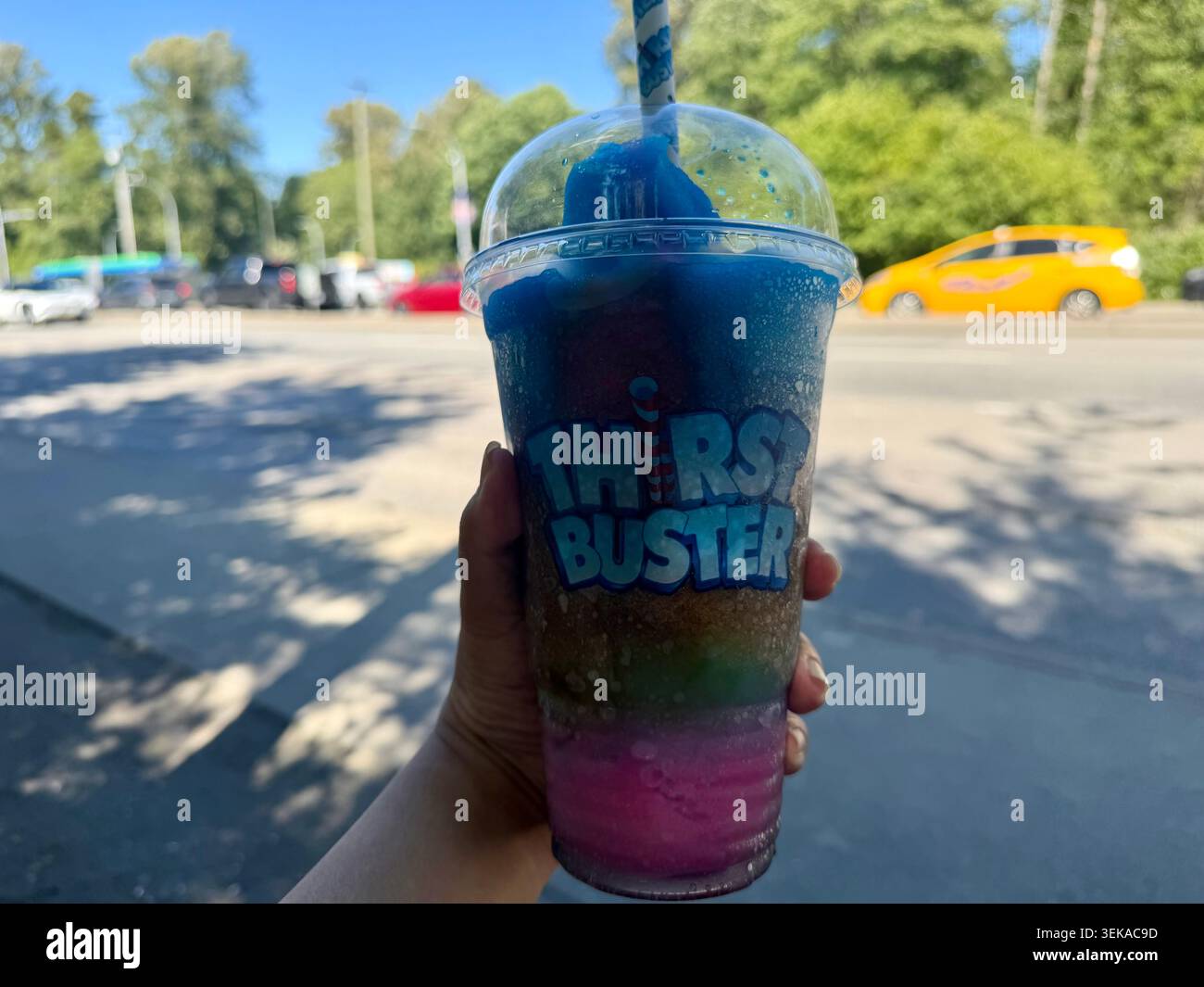 Eine Hand, die ein farbenfrohes, mehrschichtiges Slushie-Getränk in einem klaren Plastikbecher auf einem gepflasterten Fußweg hält. Stockfoto