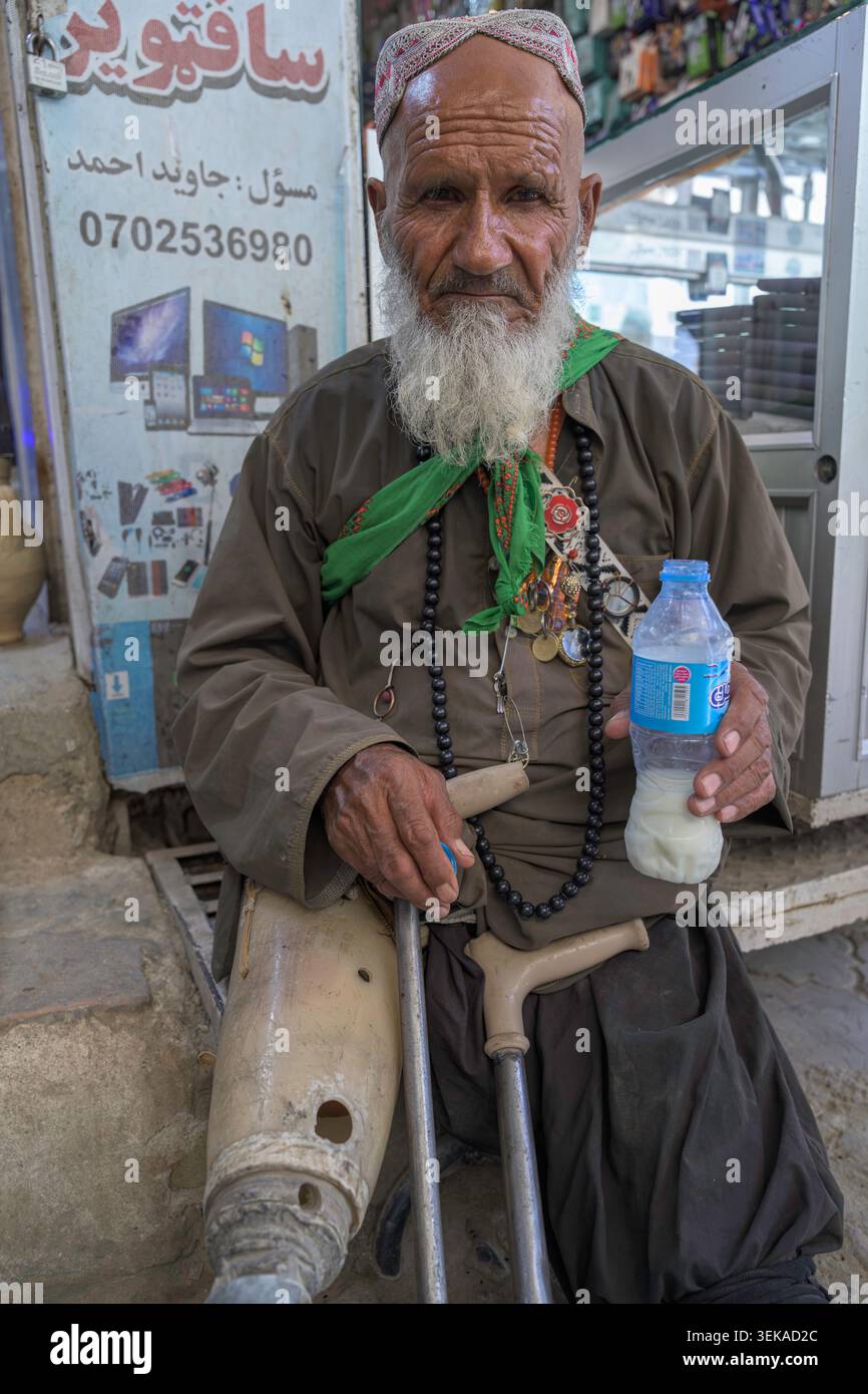 Älterer Mann mit langem weißen Bart, traditionellem Hut und Gebetskegeln, die eine Flasche Flüssigkeit halten, sitzt draußen mit Krücken und einer Beinprothese Stockfoto