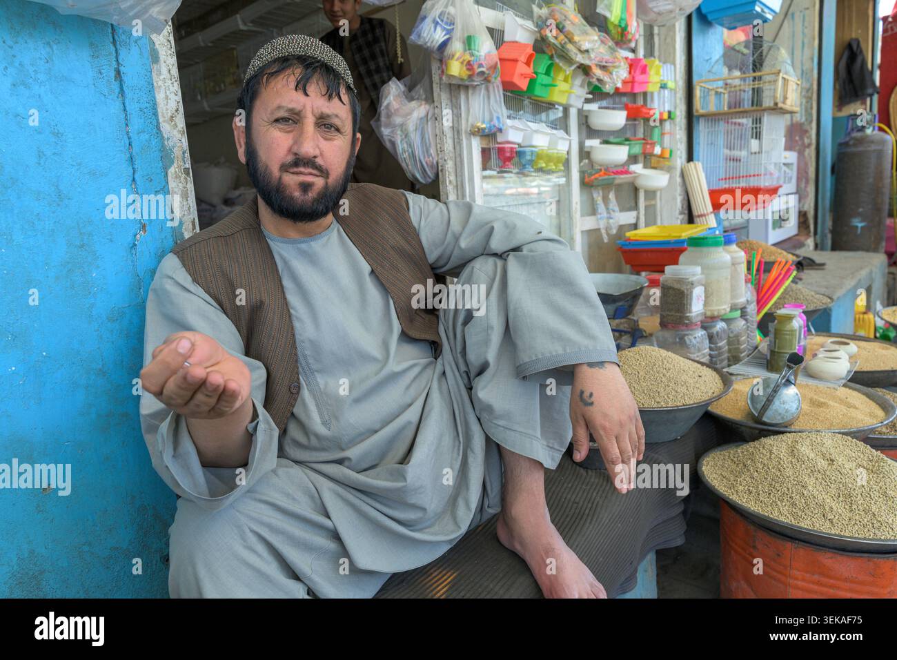 Ein Ladenbesitzer, der an seinem Marktstand sitzt und große Mengen verschiedener Getreidearten, Gewürze und Kleinartikel für Vögel zeigt. Stockfoto