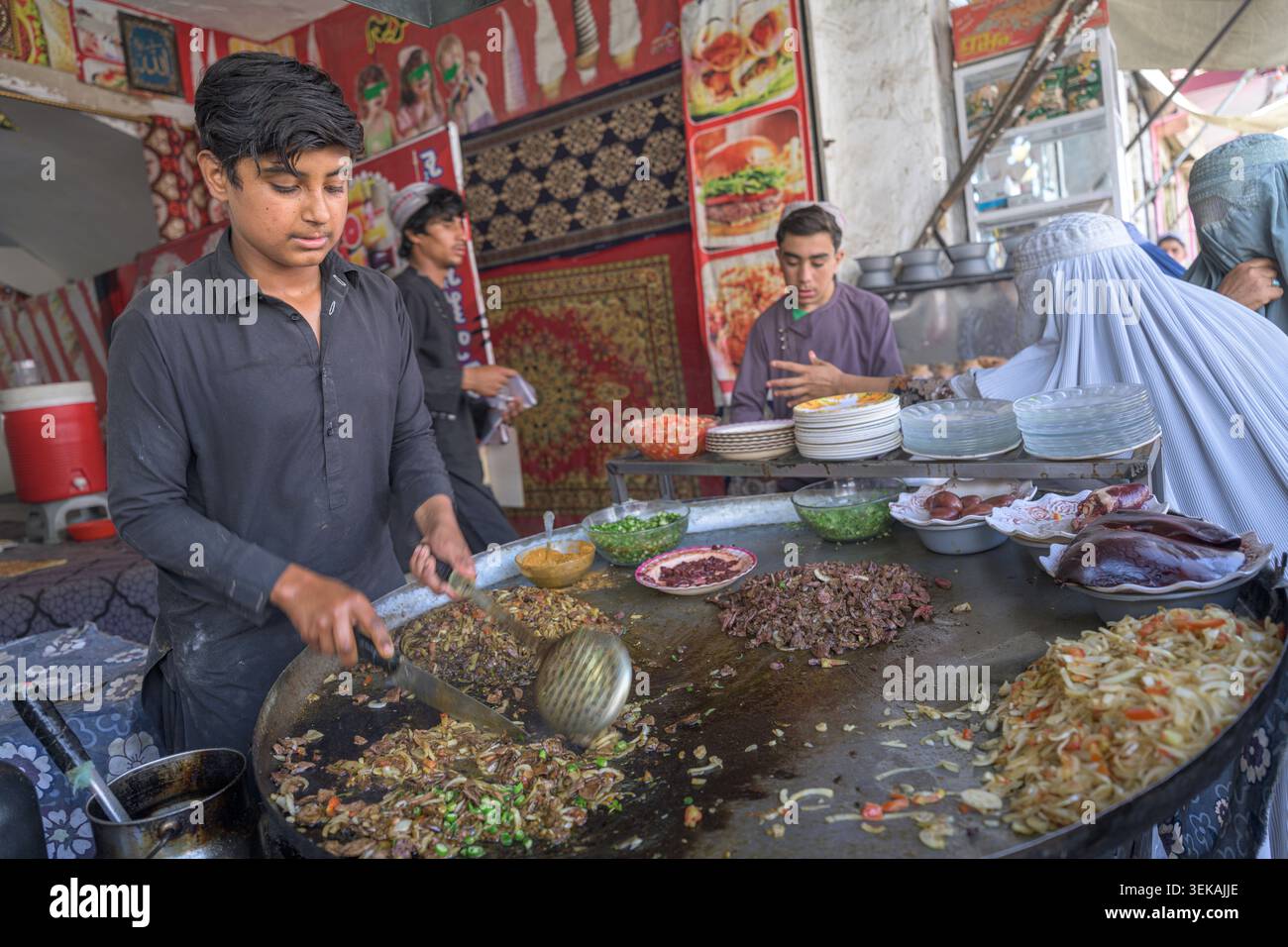 Junger Mann, der an einer großen Pfanne steht und frisches Fleisch und Gemüsegerichte für Kunden auf einem geschäftigen Straßenmarkt zubereitet. Stockfoto