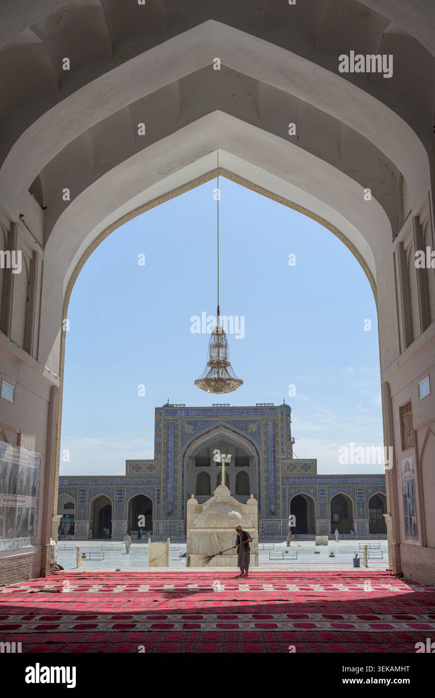 Mann, der den roten Teppich im historischen Herat großen Moschee-Innenhof fegt, eingerahmt von einem alten Bogengang. Stockfoto