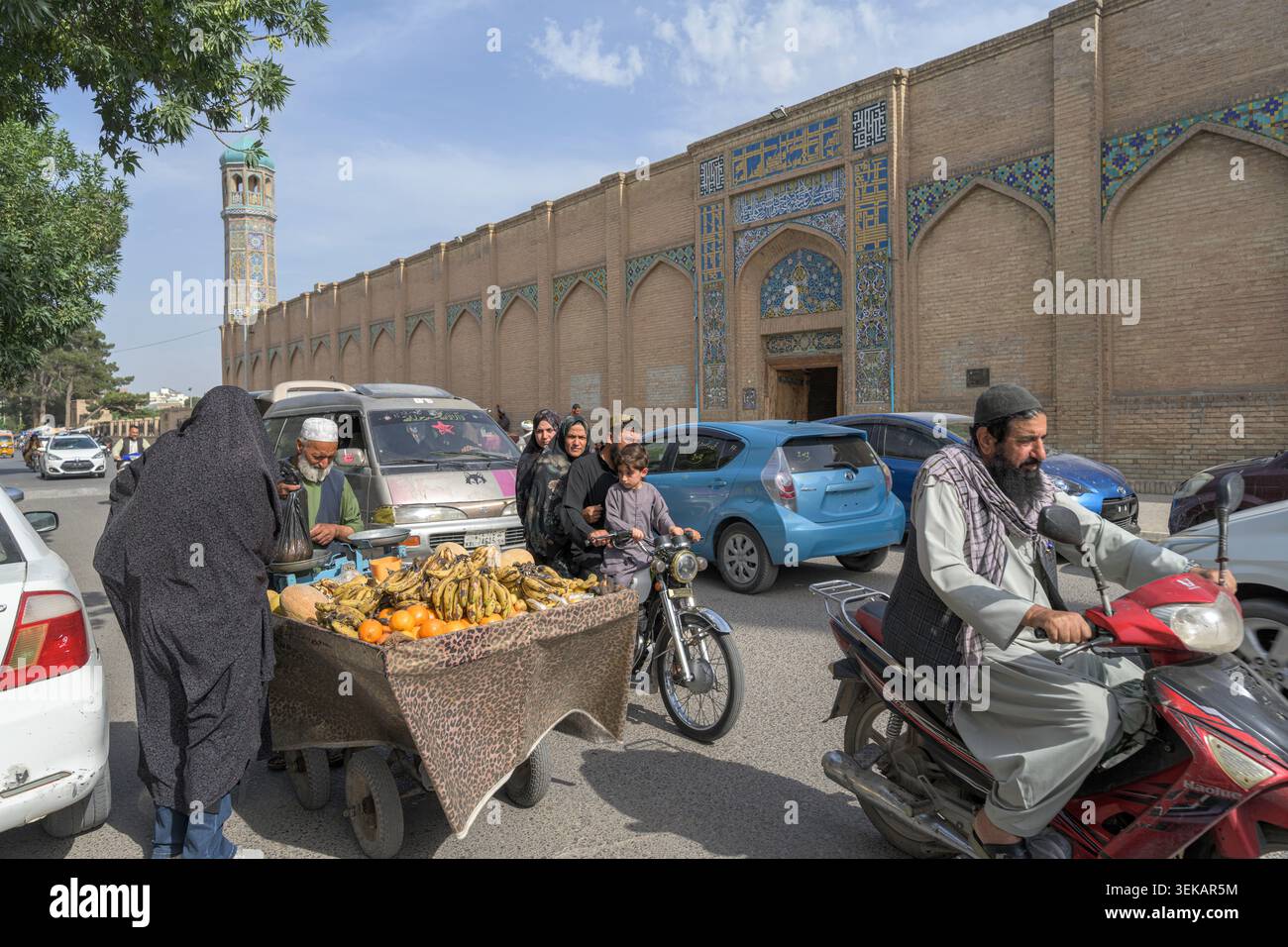 Die geschäftige Straßenszene in Herat zeigt die Aktivität des Basars mit Obstverkäufern, Menschen, Fahrzeugen und großartiger Moschee-Architektur. Stockfoto