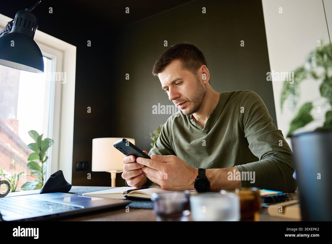 Fokussierter Mann mit Smartphone am Schreibtisch neben Laptop und offenem Notebook. Konzept der Planung, Organisation und produktiven Arbeit zu Hause. Stockfoto