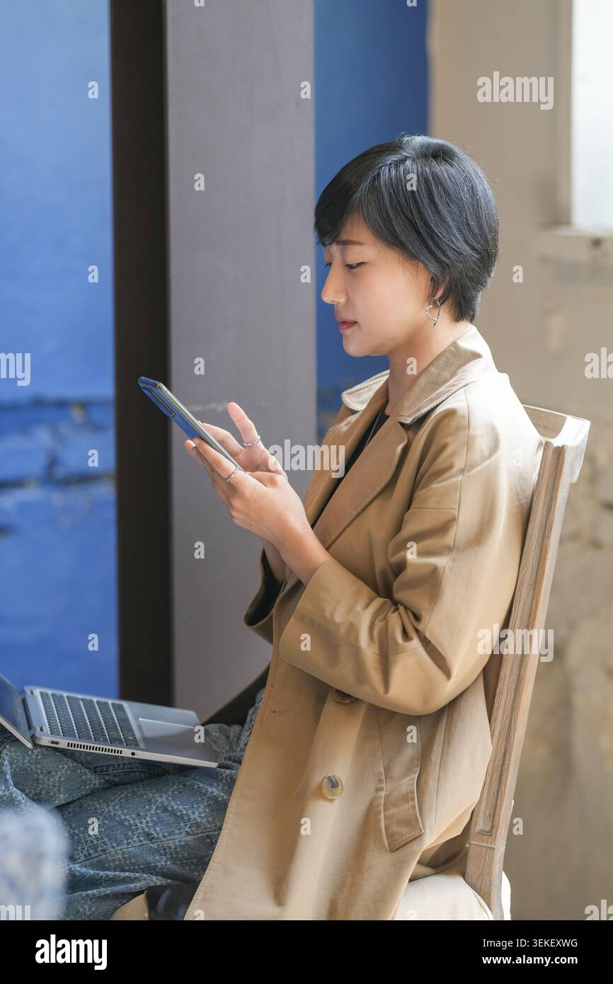 Die Frau sitzt ruhig, konzentriert auf ihr Handy. Ihr Mantel ist braun, über die Schultern gelegt. Sie beugt sich vor einer blauen Kulisse. Ihr Ausdruck ist ruhig Stockfoto