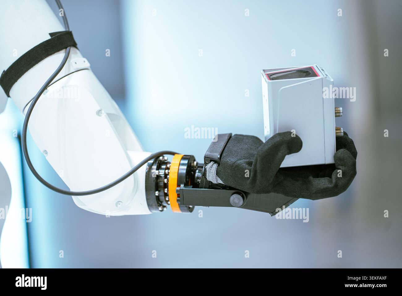 Nahaufnahme eines weißen Roboterarms mit schwarzem, mit Stoff bedecktem Endeffektor, der während einer technischen Demonstration ein graues elektronisches Bauteil hält. Stockfoto