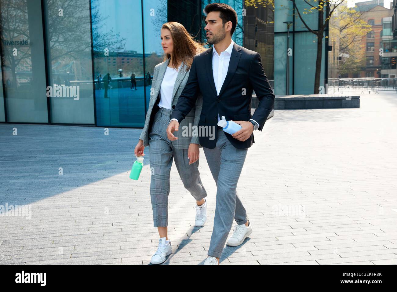 Mann und Frau in eleganten, legeren Business-Outfits, die zusammen durch einen modernen stadtplatz mit wiederverwendbaren Wasserflaschen spazieren Stockfoto