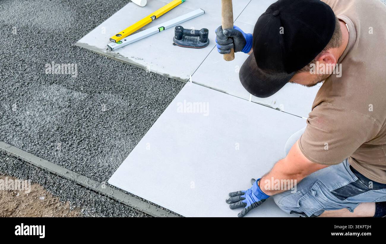 Männlicher Bauarbeiter, der Betonziegel mit Hammer und Nivellierwerkzeug auf Kies legt, mit zusätzlichen Werkzeugen, die im Hintergrund sichtbar sind Stockfoto