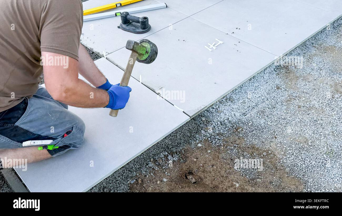 Männlicher Arbeiter, der große Betonziegel mit einem Gummihammer, im Baubereich sichtbaren Werkzeugen und Messgeräten auf Kies legt Stockfoto