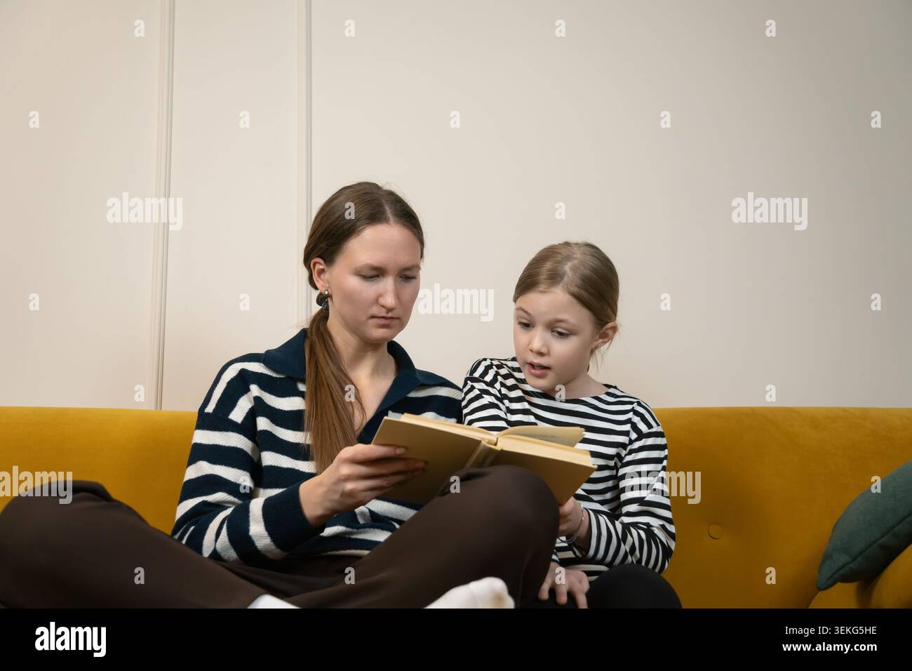 Mutter und Tochter verbinden sich beim Lesen eines Buches, sitzen zusammen auf einem gelben Sofa zu Hause, nehmen an einem friedlichen gemeinsamen Lernerlebnis Teil und enjo Stockfoto