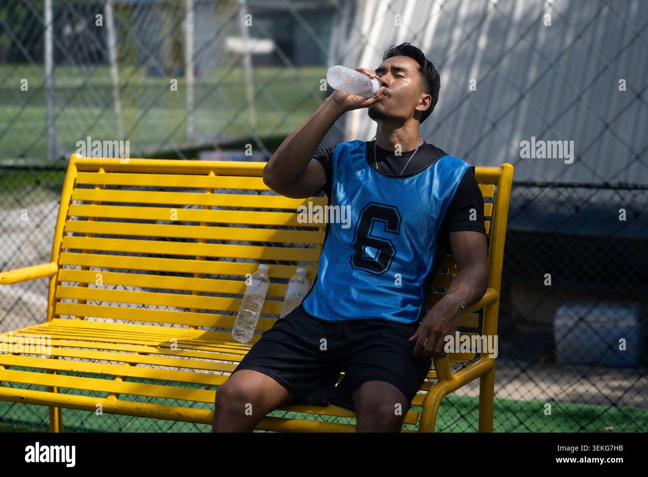 Ein thailändischer asiatischer Fußballspieler trinkt aus einer Wasserflasche, während er sich während einer Pause auf einer gelben Bank neben einem Außenfeld ausruht Stockfoto