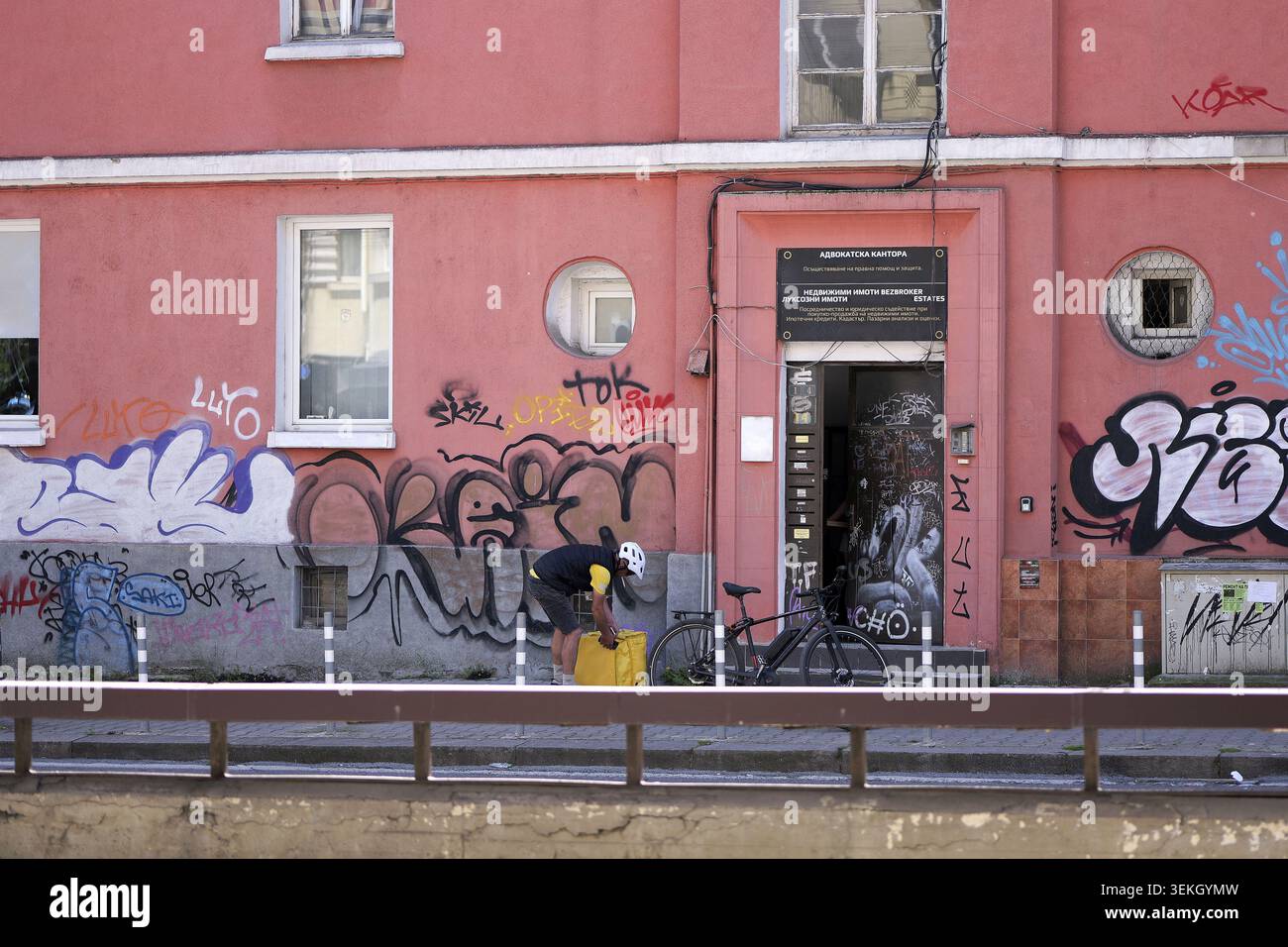 Lieferer in Schutzhelm und Uniform mit seinem Fahrrad, der eine gelbe Thermotasche vor einem Gebäude mit abblätternder Farbe, Graffiti, A sichert Stockfoto