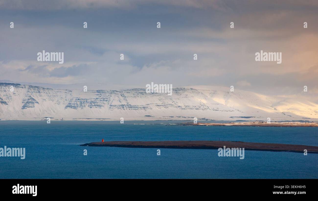 Blaues Wasser, schmale Insel mit orangefarbener Struktur. Schneebedeckte Berge unter bewölktem Himmel, Sonnenlicht auf der rechten Seite. Stockfoto