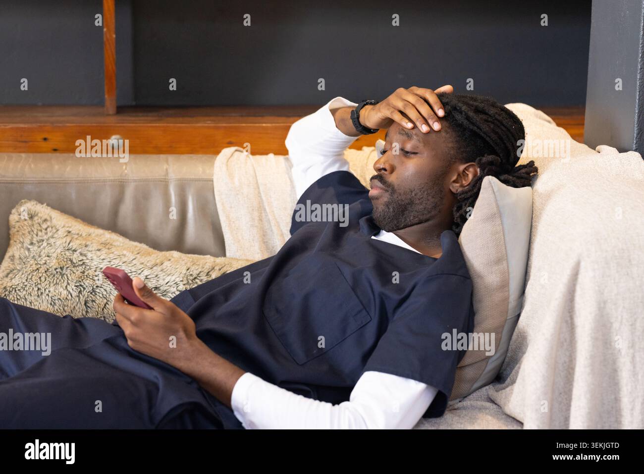 Afrikanischer Mann, der sich auf einem beigen Sofa in der Lounge liegend, mit marineblauen medizinischen Peelings, die ein rotes Smartphone halten. Couch, blau, Telefon, kontemplativ, Ermüdung, Innenraum Stockfoto