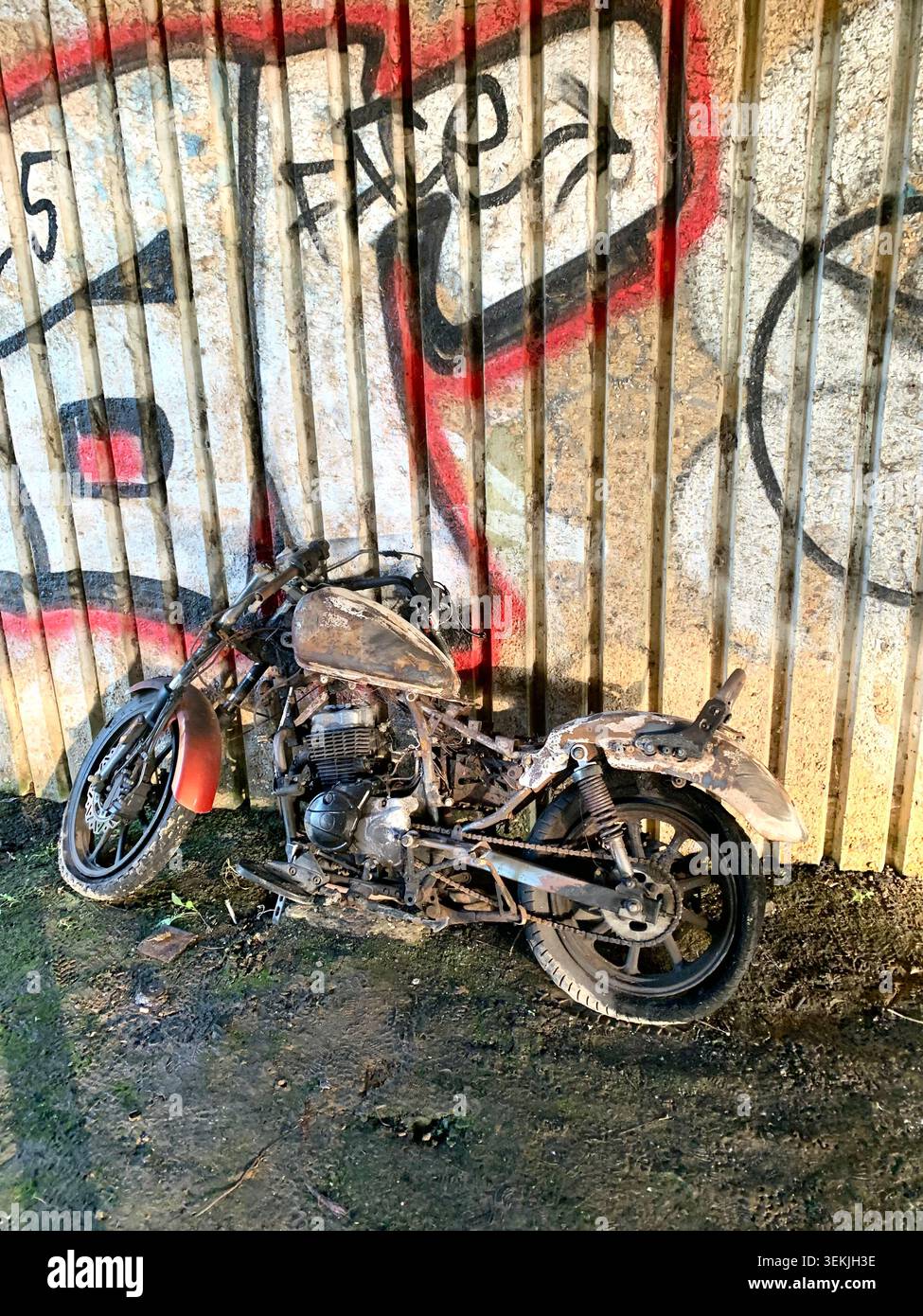 Verlassenes Motorrad unter einer mit Graffiti bedeckten Betonmauer Northamptonshire England verrostete Rahmen und fehlende Teile erinnern an urbanen Verfall - Smartphone-aufgenommenes Stockfoto