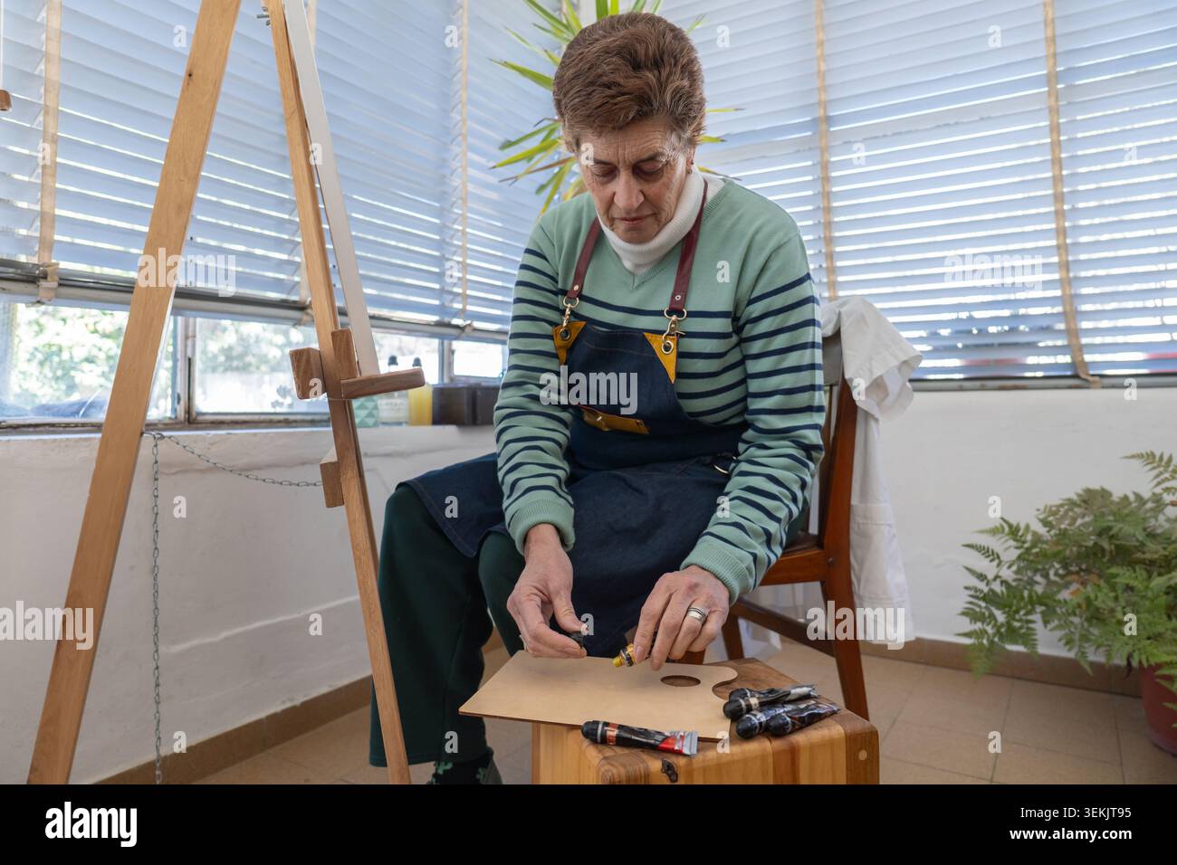 Eine reife Künstlerin in einer Denim-Schürze sitzt auf einem Stuhl neben ihrer hölzernen Staffelei und drückt Farbe aus einem Rohr auf eine Holzpalette, die auf einem kleinen s liegt Stockfoto