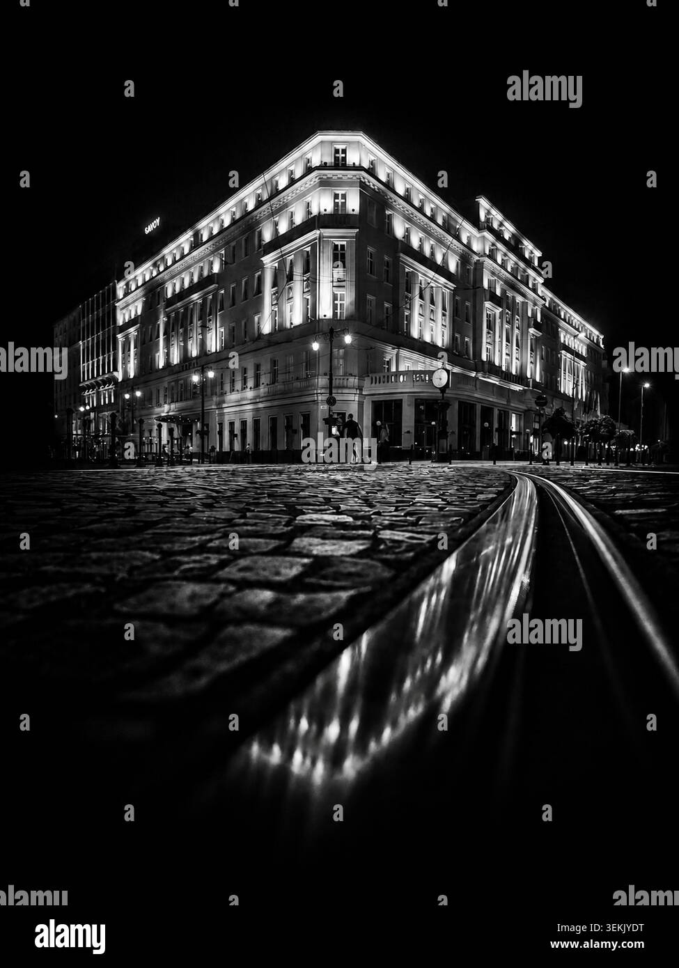 Schwarz-weiße Nachtansicht auf das Carlton Hotel in Bratislava, Slowakei, mit leuchtender Fassade, Kopfsteinpflaster und Straßenbahnlinien. Stockfoto