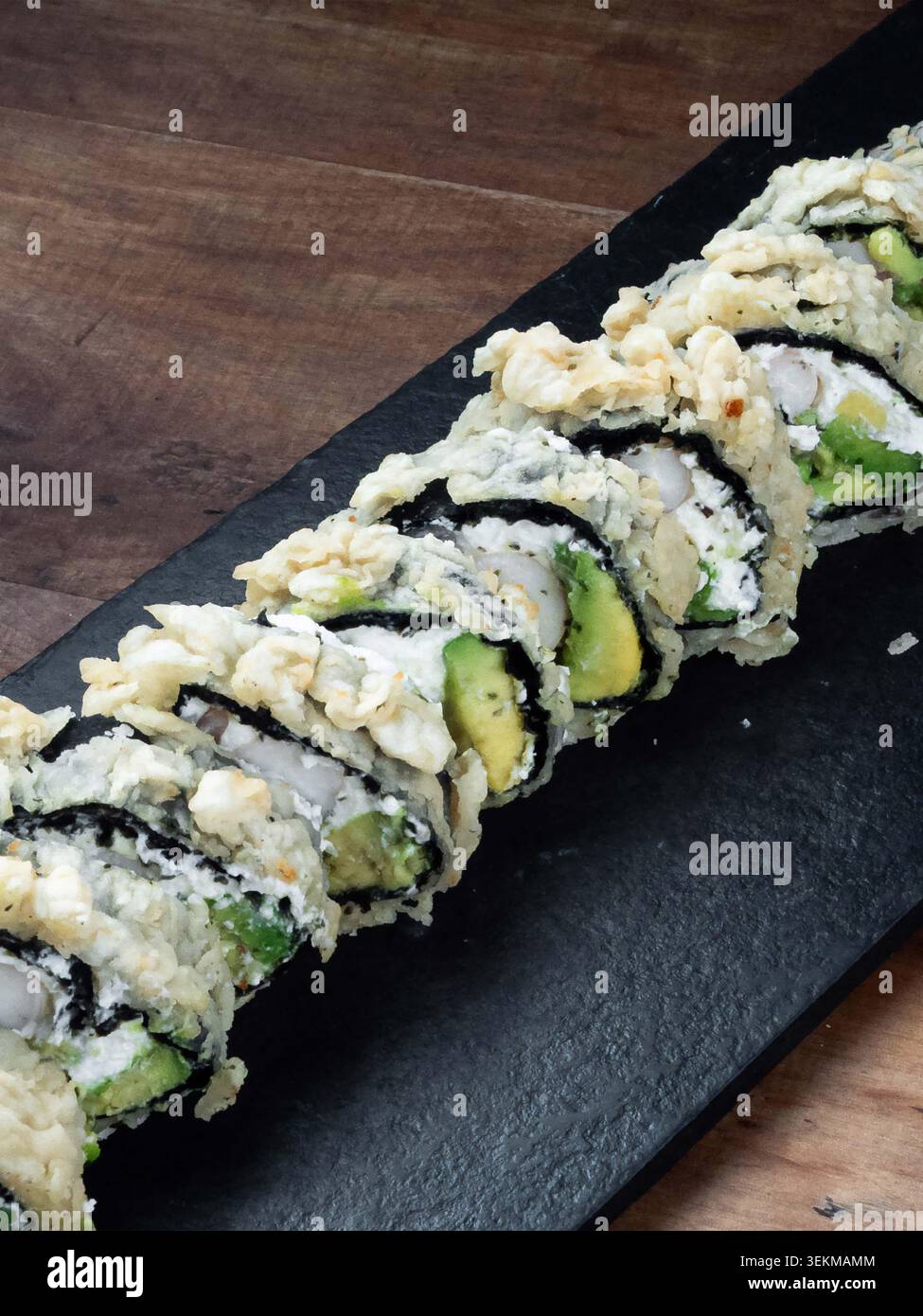 Knuspriger, goldbrauner Teig umhüllt jede Sushi-Rolle. Avocado und Weißfisch füllen die Brötchen ordentlich. Sushi liegt auf einem dunklen, eleganten Servierteller. Hölzerner Su Stockfoto