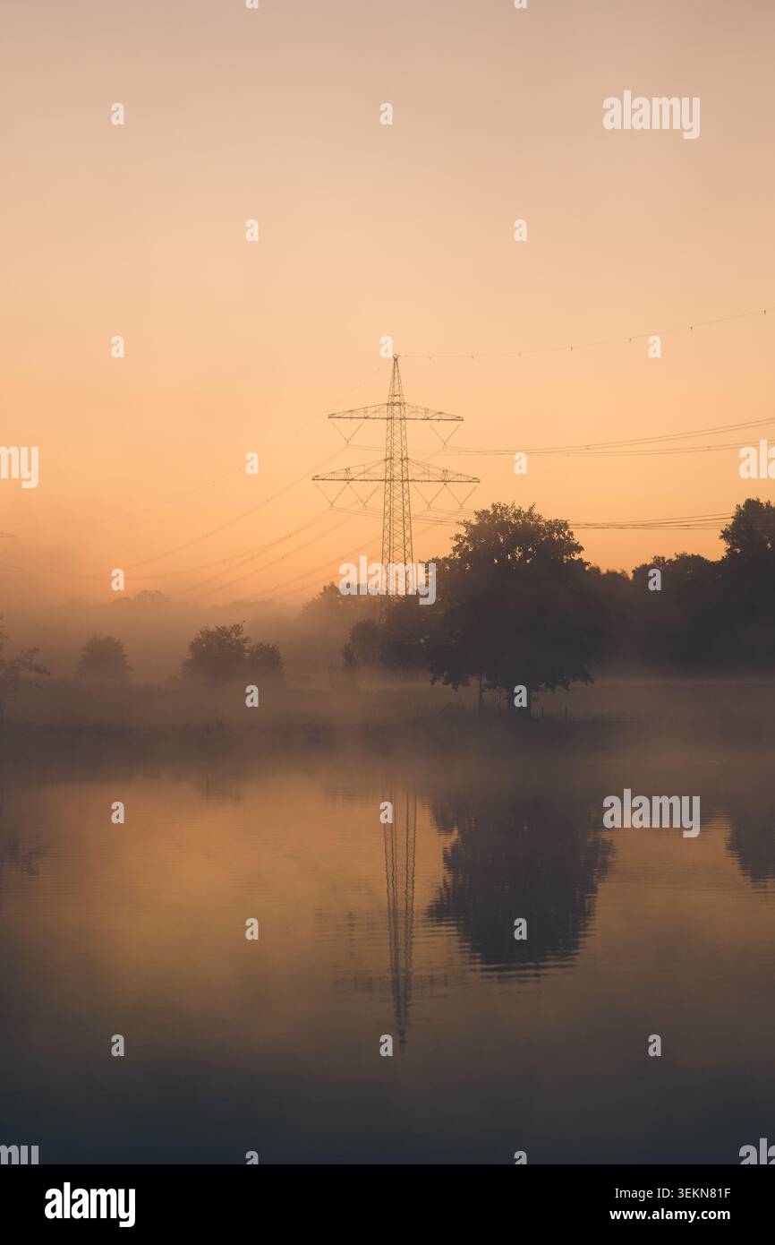 Über einem ruhigen, ländlichen See steht bei Sonnenaufgang ein hoher Strompylon mit weichem Nebel, Silhouettenbäumen und warmem goldenem Licht, das auf dem Wasser reflektiert wird Stockfoto