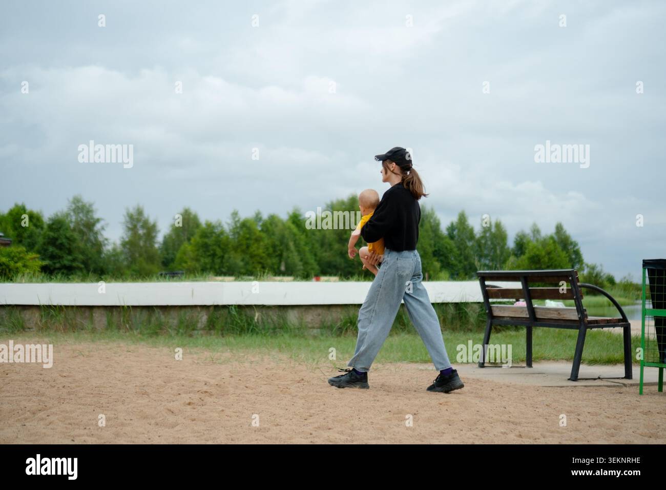 Eine pflegende Frau hält ihr Kind nahe, wenn sie durch einen Park läuft. Die schützende Mutter trägt das Kleinkind sanft auf einem sandigen Pfad im Freien. Liebevolle Eltern und Baby teilen einen zärtlichen Moment zwischen Bäumen Stockfoto