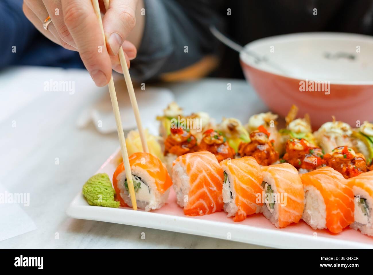 Eine Handperson hält Essstäbchen über einem Teller mit frischen Sushi-Brötchen in einem Restaurant. Diner wählt ein Stück Lachsnigiri aus der Sorte. Asiatische Küche mit Suppenschüssel im Hintergrund Stockfoto