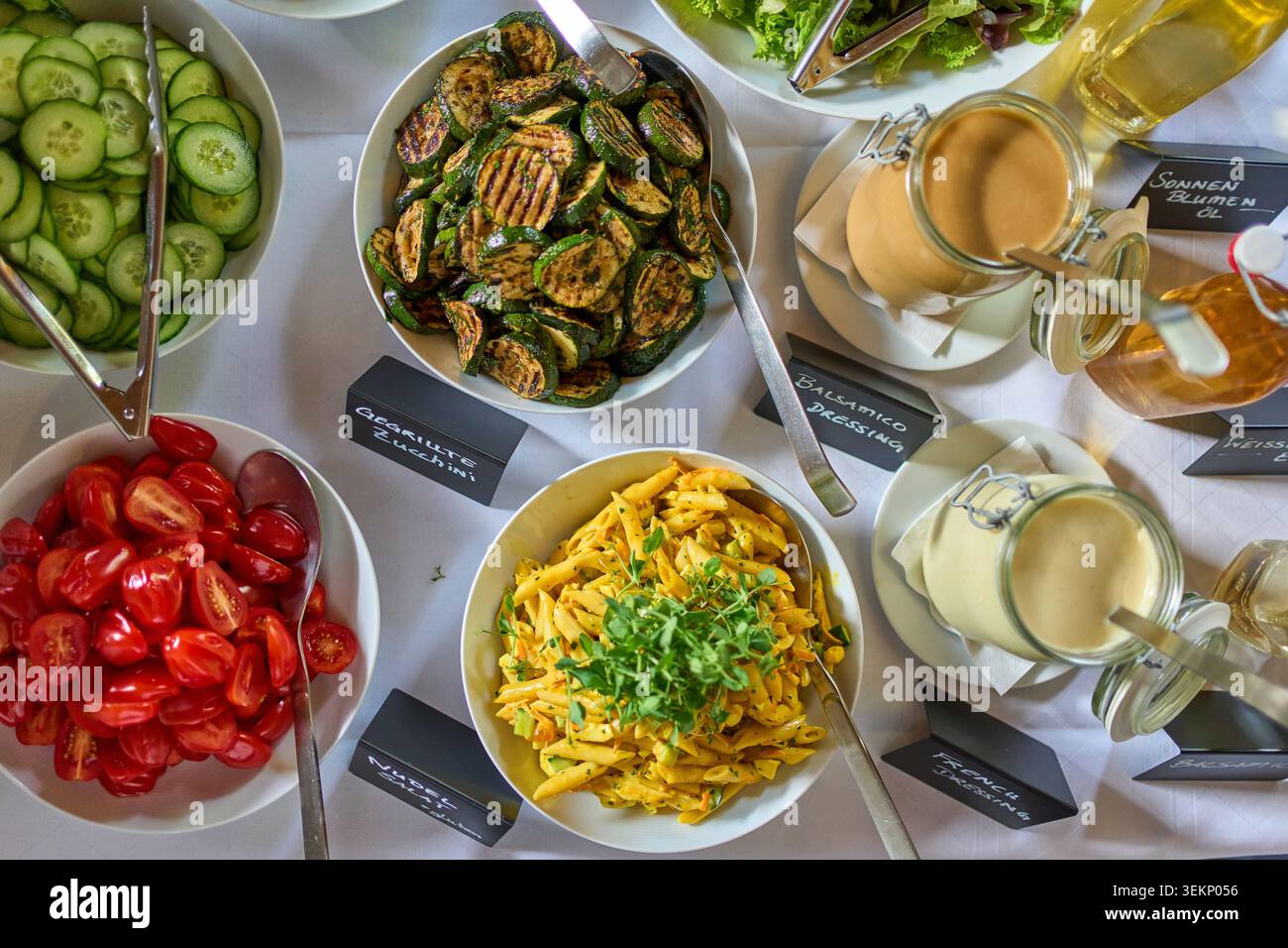 Ein Catering-Buffet mit Blick auf die Spitze mit weißen Schüsseln mit Gurken, Tomaten, gegrillten Zucchini, Nudelsalat, Dressings, und Öle auf einer weißen Tischdecke. Stockfoto