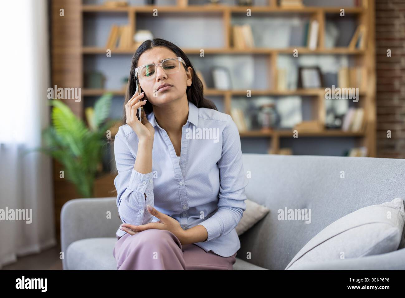 Eine gestresste junge Frau mit Brille fühlt sich müde und frustriert, hat Kopfschmerzen, während sie auf einer Couch sitzt und während eines schwierigen Telefonanrufs zu Hause auf einem Smartphone spricht Stockfoto