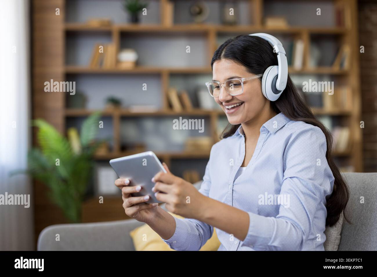 Junge Frau mit Brille und Kopfhörern, die sich auf dem Sofa entspannt und lächelt, während sie zu Hause Videos und Musik auf einem Tablet streamt, um Unterhaltung, Lernen oder E-Learning zu genießen Stockfoto
