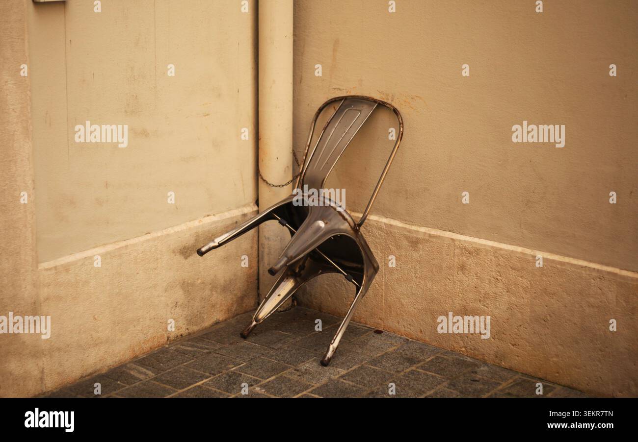 Ein einzelner, abgenutzter Metallstuhl, der sich unangenehm in einer Gebäudeecke beugte und fest mit einer Kette an einem alten vertikalen Abflussrohr befestigt wurde. Stockfoto