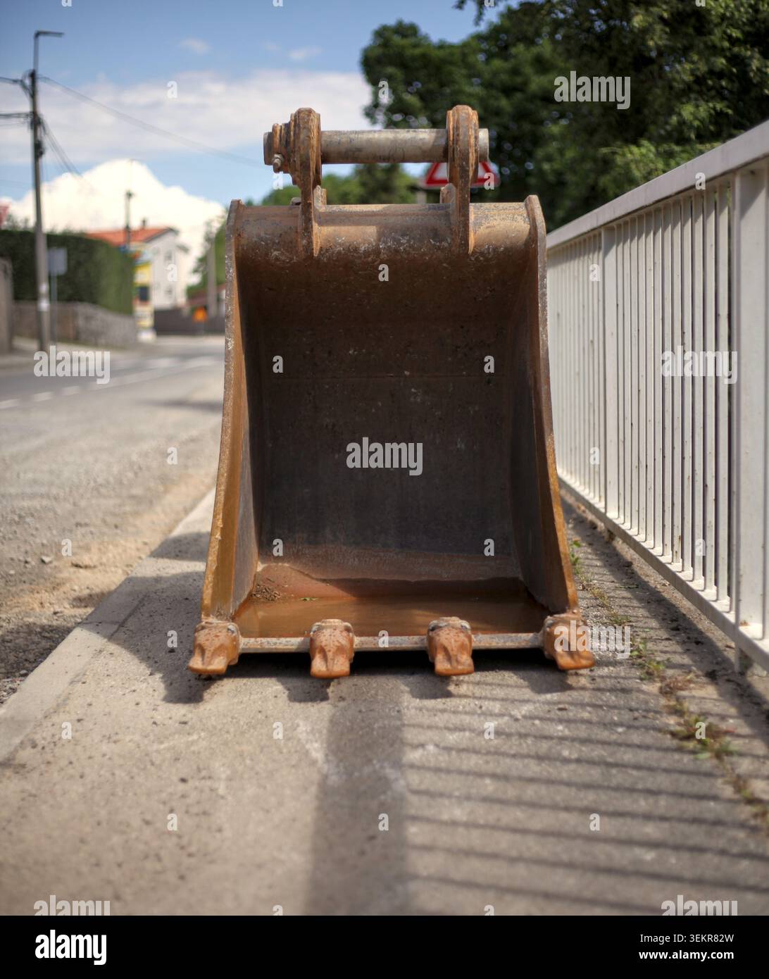 Ein leerer, rostiger Baggerlöffel, der auf einem Gehsteig aus Beton neben einem weißen Metallzaun an einer Straße sitzt. Stockfoto