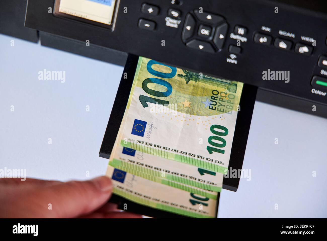 Nahaufnahme einer menschlichen Hand, die gedruckte 100-Euro-Banknoten aus einem Inkjet-Druckerfach für das Heimbüro aufnimmt, um das Konzept der Fälschung zu veranschaulichen. Stockfoto