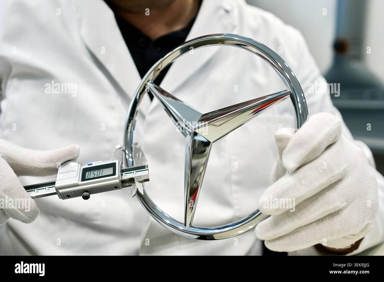Ein Inspektor zur Qualitätskontrolle mit einem digitalen Messschieber, der die genauen Maße eines verchromten Mercedes-Benz Stern-Logos in einem Automobilwerk überprüft. Stockfoto