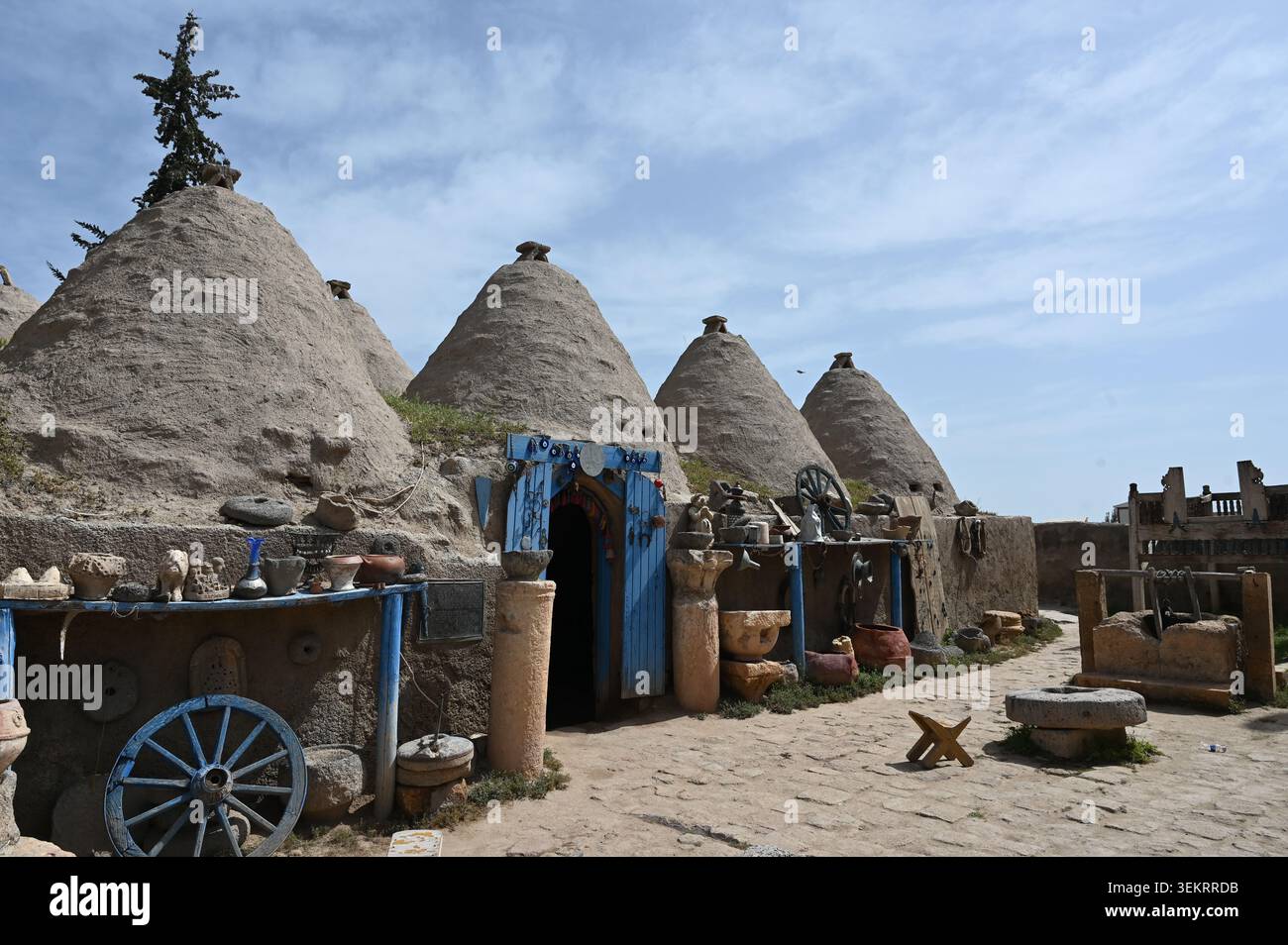 Traditionelle Bienenstockhäuser und Handwerksstand in Harran, Şanlıurfa, Türkei. Erdige kegelförmige Behausungen, blaue Tür und Töpferwaren unter einem hellblauen Himmel Stockfoto