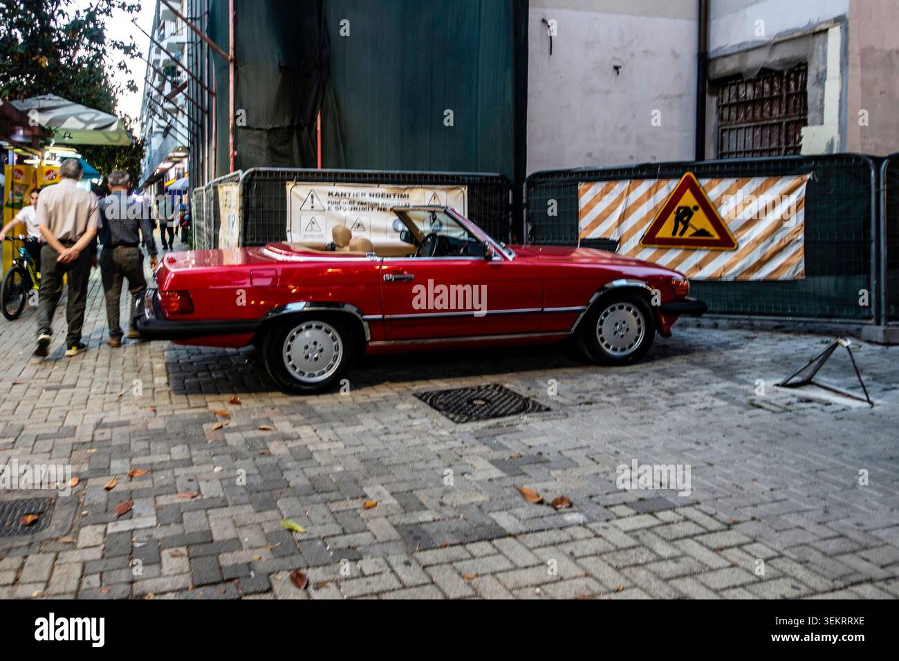 Ein rotes Mercedes-Benz SL Cabriolet steht auf einem Backsteinpflaster in der Nähe einer umzäunten Baustelle, an der Fußgänger in Tirana, Alba, vorbeilaufen Stockfoto