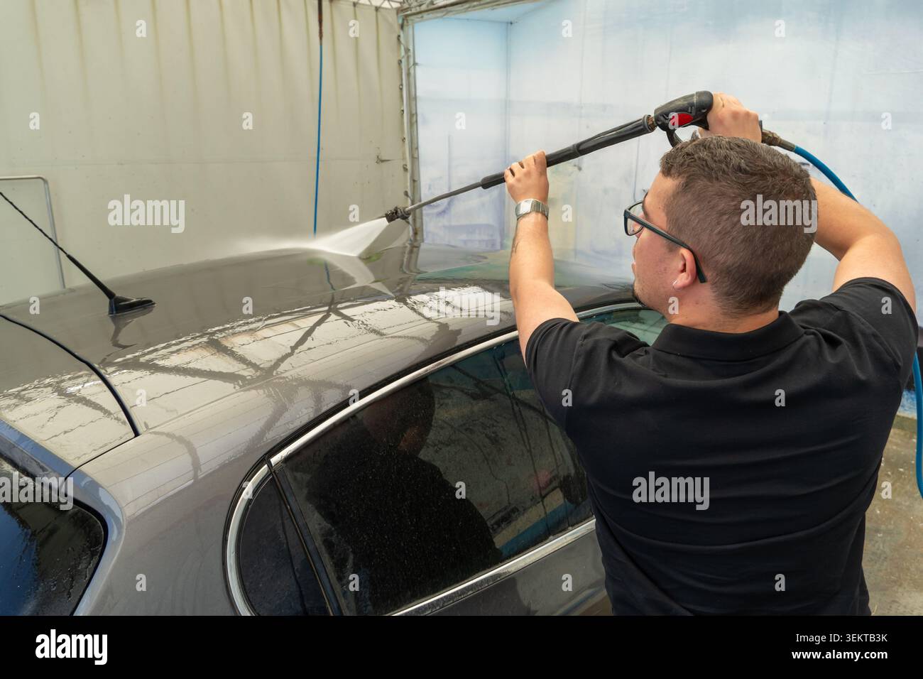 Mann, der das Dach eines grauen Fahrzeugs mit einem Hochdruckreiniger in einer automatischen Waschanlage reinigt und reinigt Stockfoto