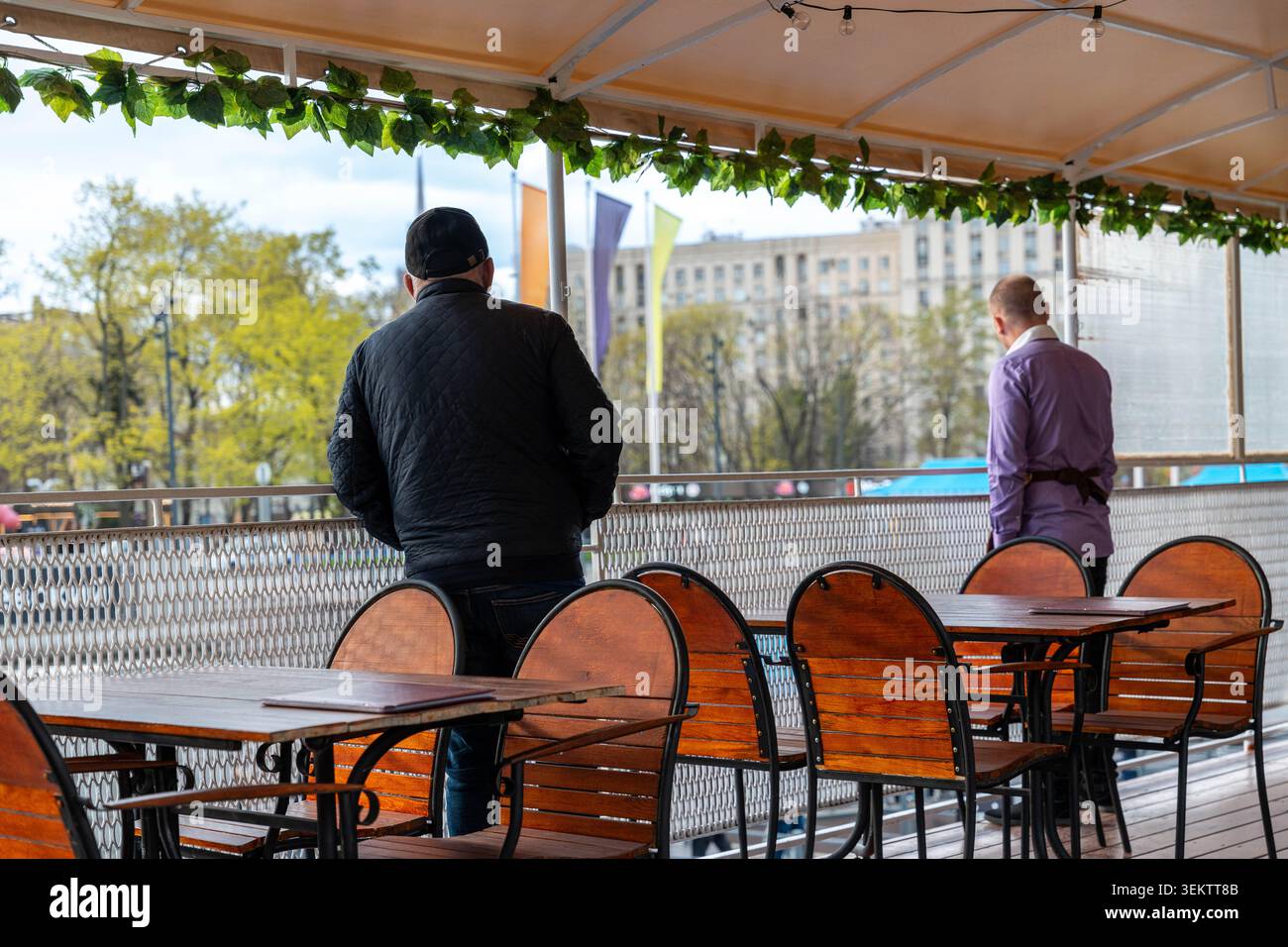 Zwei Männer Auf Der Riverside Terrace Mit Blick Auf Die Skyline Der Stadt, Am Geländer Neben Leeren Cafe-Tischen Im Herbstlicht, Lässigen Mänteln Und Cap, Ruhig Stockfoto