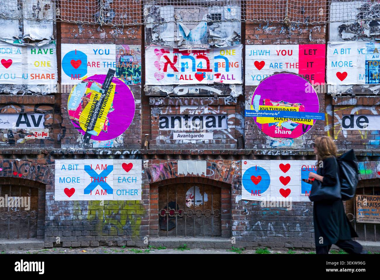 Fußgänger, der an einer verwitterten Ziegelwand vorbeigeht, die mit Plakaten, Street Art und Typografie mit dem Wort „Avantgarde“ bedeckt ist. Stockfoto