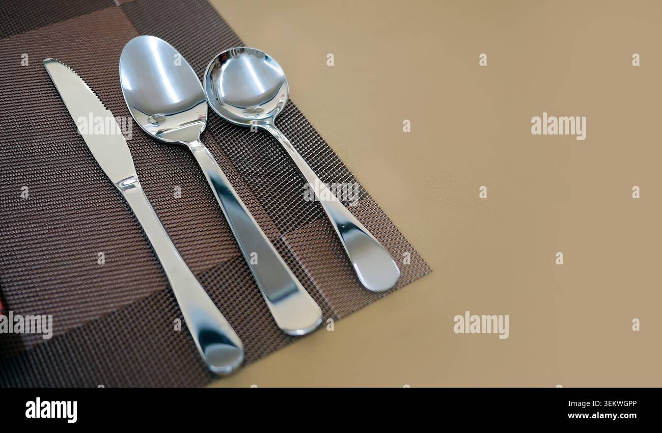 Poliertes Bestecktrio aus Edelstahl (Messer, Esslöffel und Suppenlöffel), ordentlich auf einem dunkelbraunen, gewebten Tischset angeordnet. Hochwinkelaufnahme mit smo Stockfoto