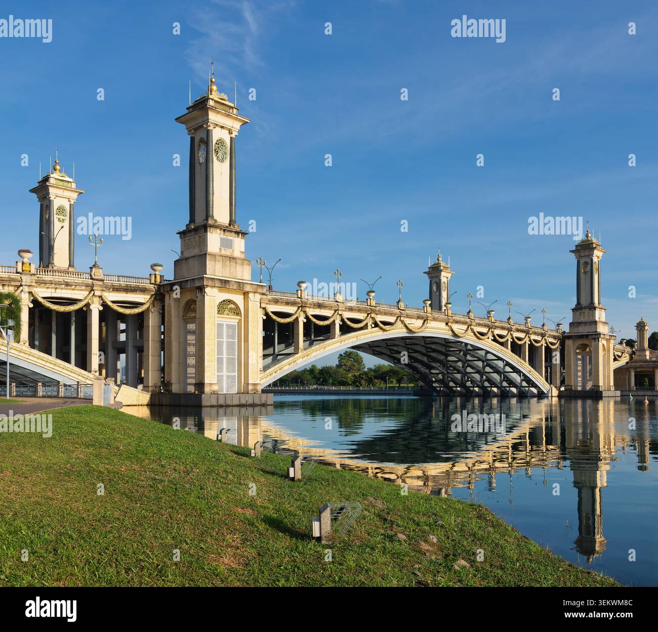 Die reich verzierte Seri Gemilang Brücke in Putrajaya, Malaysia, bietet große Türme und goldene Verzierungen, die sich wunderbar im ruhigen Wasser unter einem klaren blauen Himmel spiegeln Stockfoto