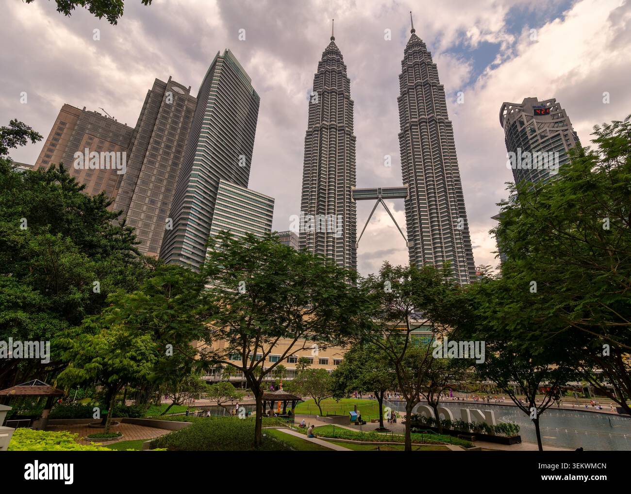 Kuala Lumpur, Malaysia - 17. Februar 2025: Die legendären Petronas Twin Towers erheben sich über üppige Bäume und moderne Gebäude unter einem bewölkten Himmel, mit einem Park und einem Brunnen darunter Stockfoto