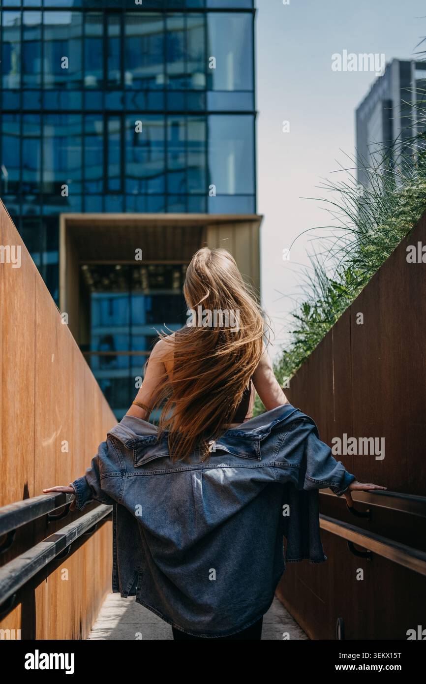 Frau geht die Fußgängerrampe zwischen Wänden hinauf und blickt auf moderne Glashochhäuser. Begehbare Stadtinfrastruktur, städtischer Fußgängerweg, Smart Stockfoto